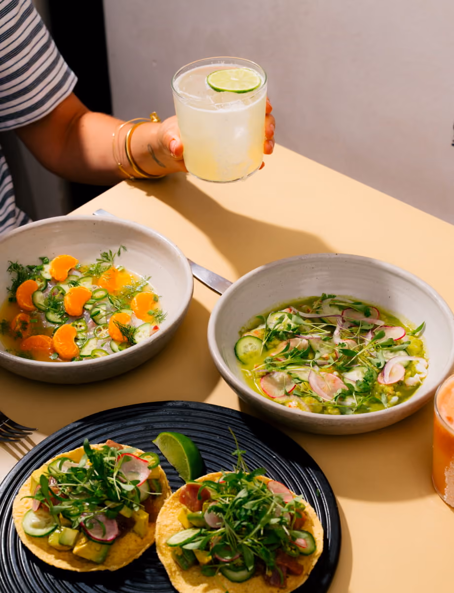 Mexican-inspired spread featuring tostadas, ceviche bowls with citrus and radish, and a hand holding a lime margarita