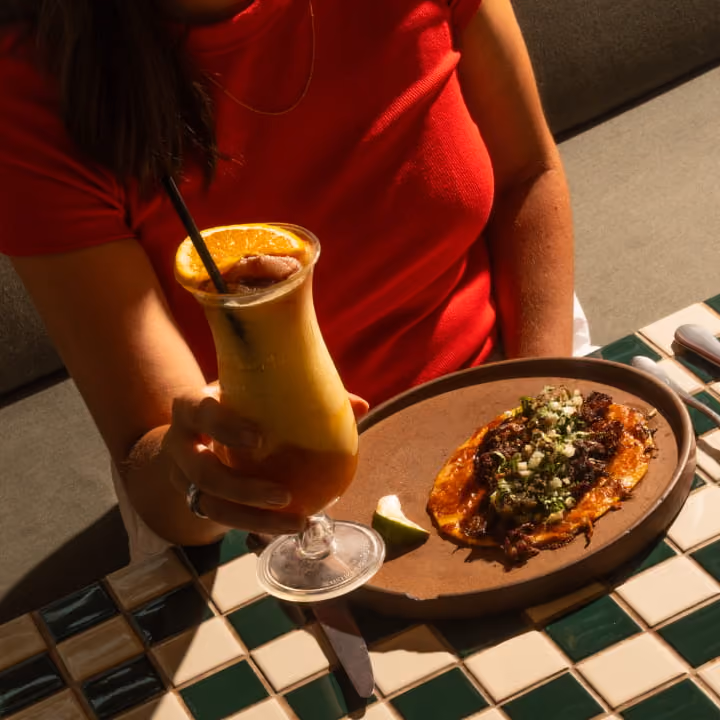 Woman in a red top holding a frozen orange cocktail beside a birria taco on a brown ceramic plate at a tiled table.