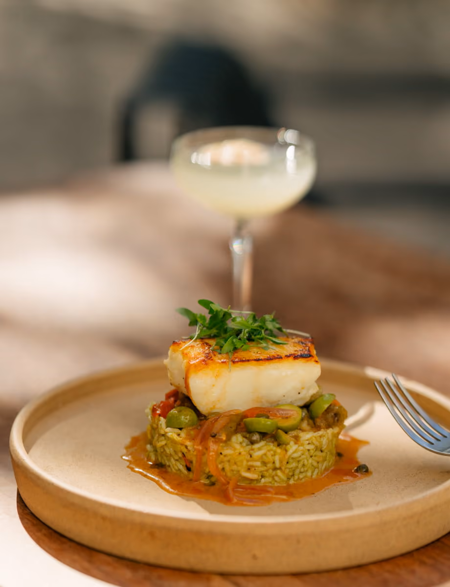 Pan-seared white fish fillet served over seasoned rice with olives and sauce, paired with a coupe cocktail glass