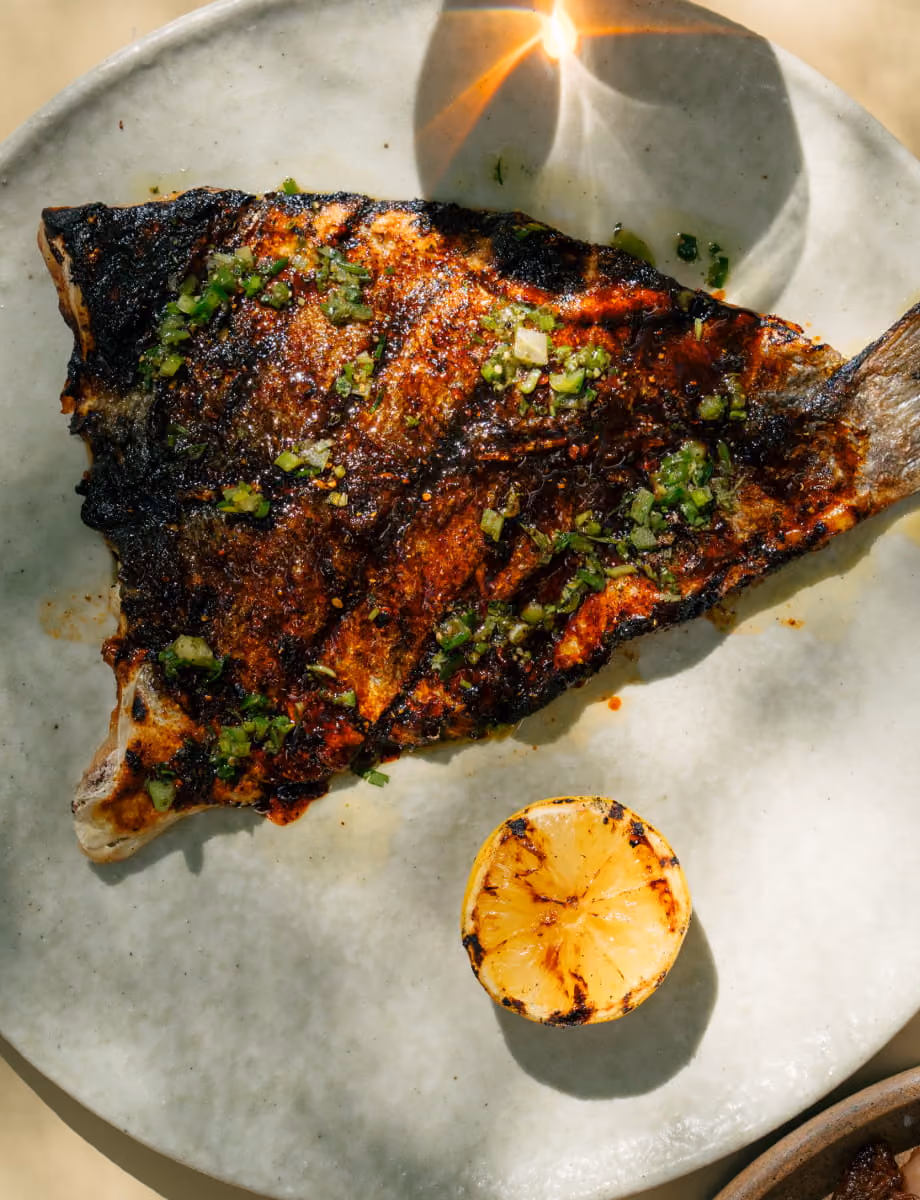 Grilled whole fish with charred skin, topped with green herb salsa and served with a grilled lemon half on a ceramic plate.
