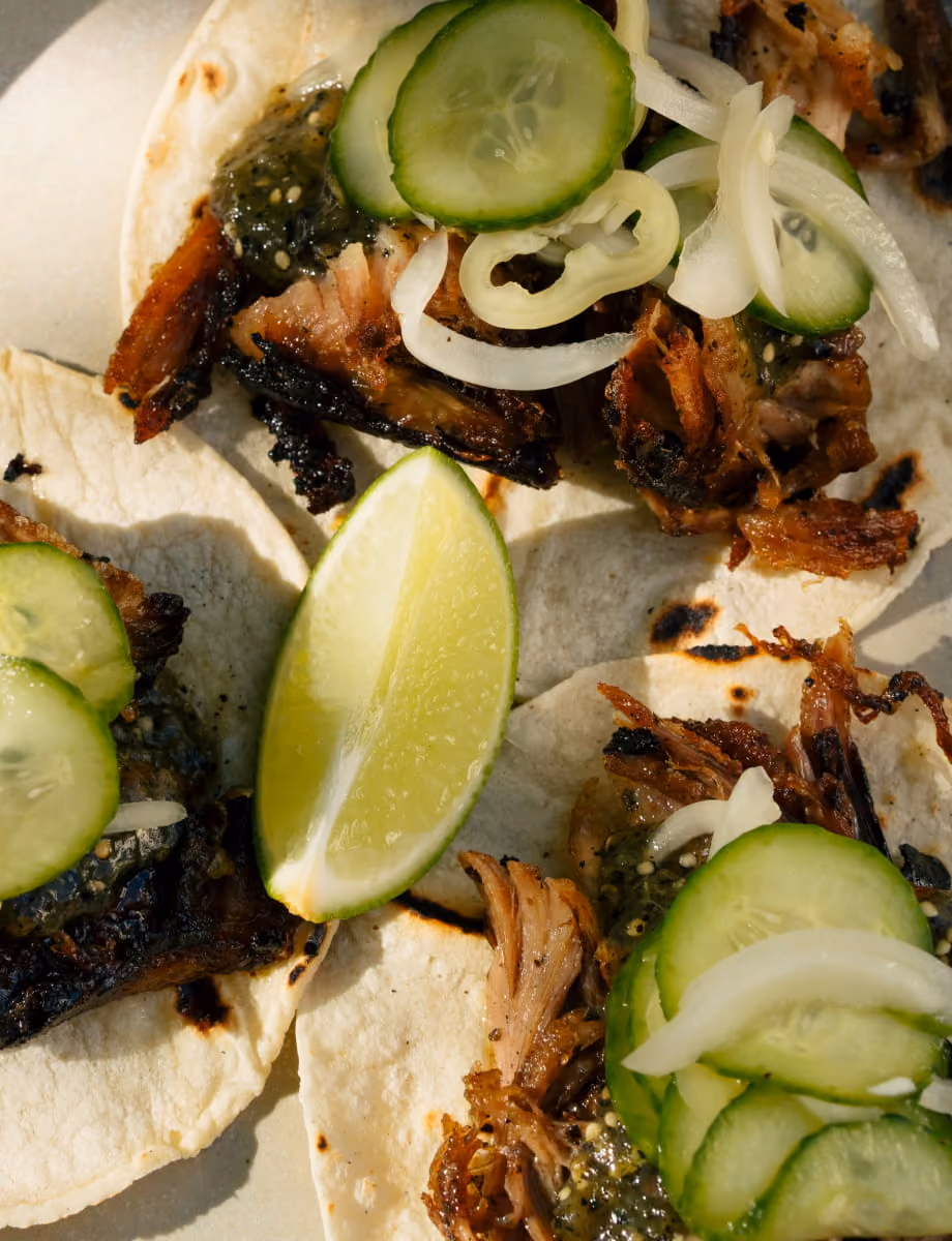 Close-up of slow-roasted pork carnitas tacos on charred flour tortillas with cucumber, onion, and fresh lime wedges