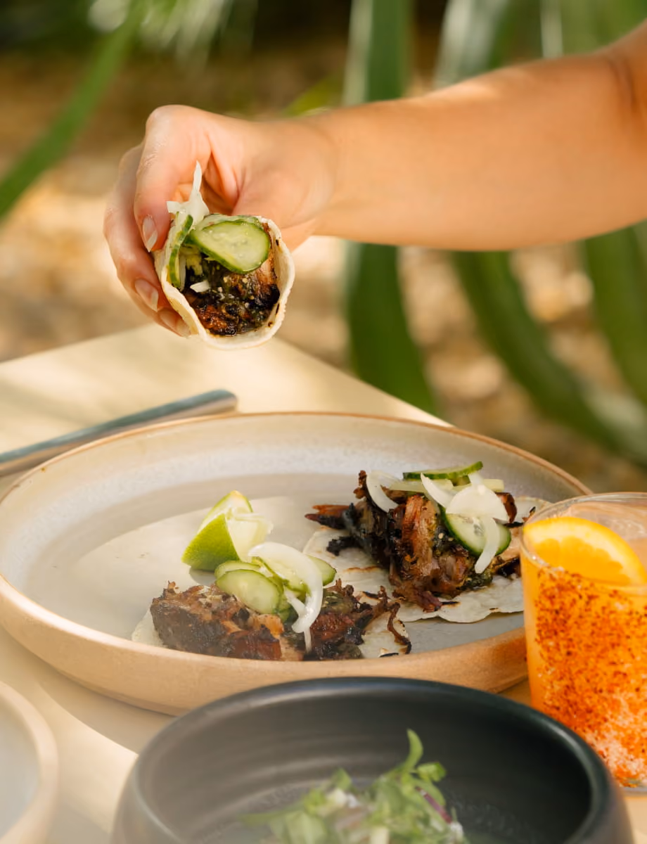 Hand holding a carnitas taco topped with cucumber and onion, served alongside a chili-rimmed michelada cocktail