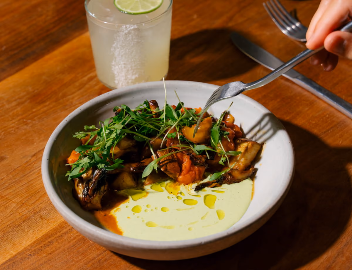 Roasted eggplant dish with tomatoes, fresh herbs, and green cream sauce on a white bowl beside a lime cocktail.