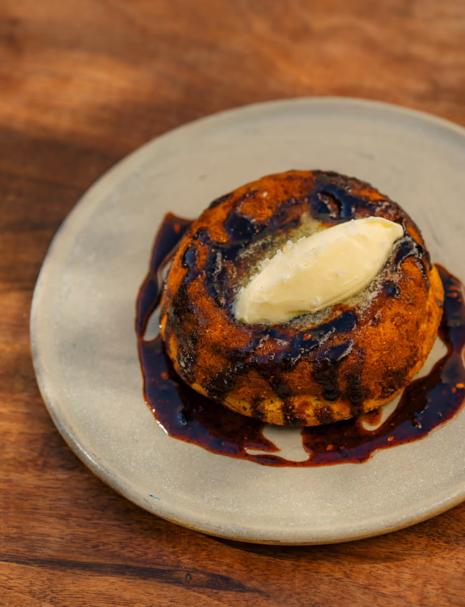 Warm cornbread topped with a melting pat of butter and drizzled with dark chile-infused sauce on a stoneware plate.