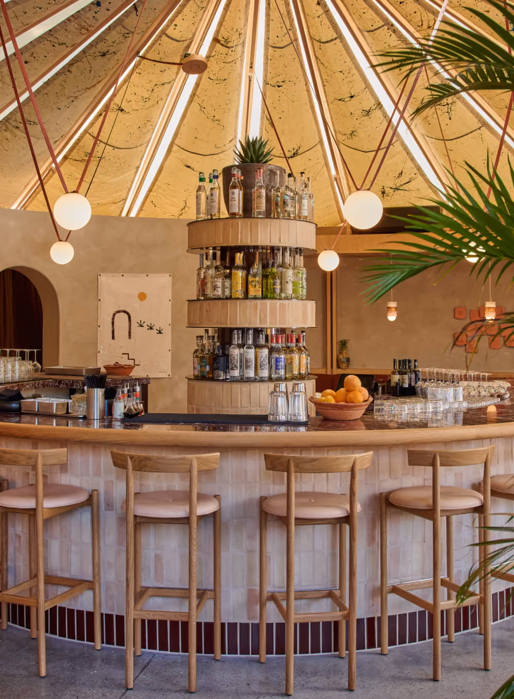 Curved tiled bar with wooden stools beneath a dramatic tent-like conical ceiling with tiered bottle display and globe lights.