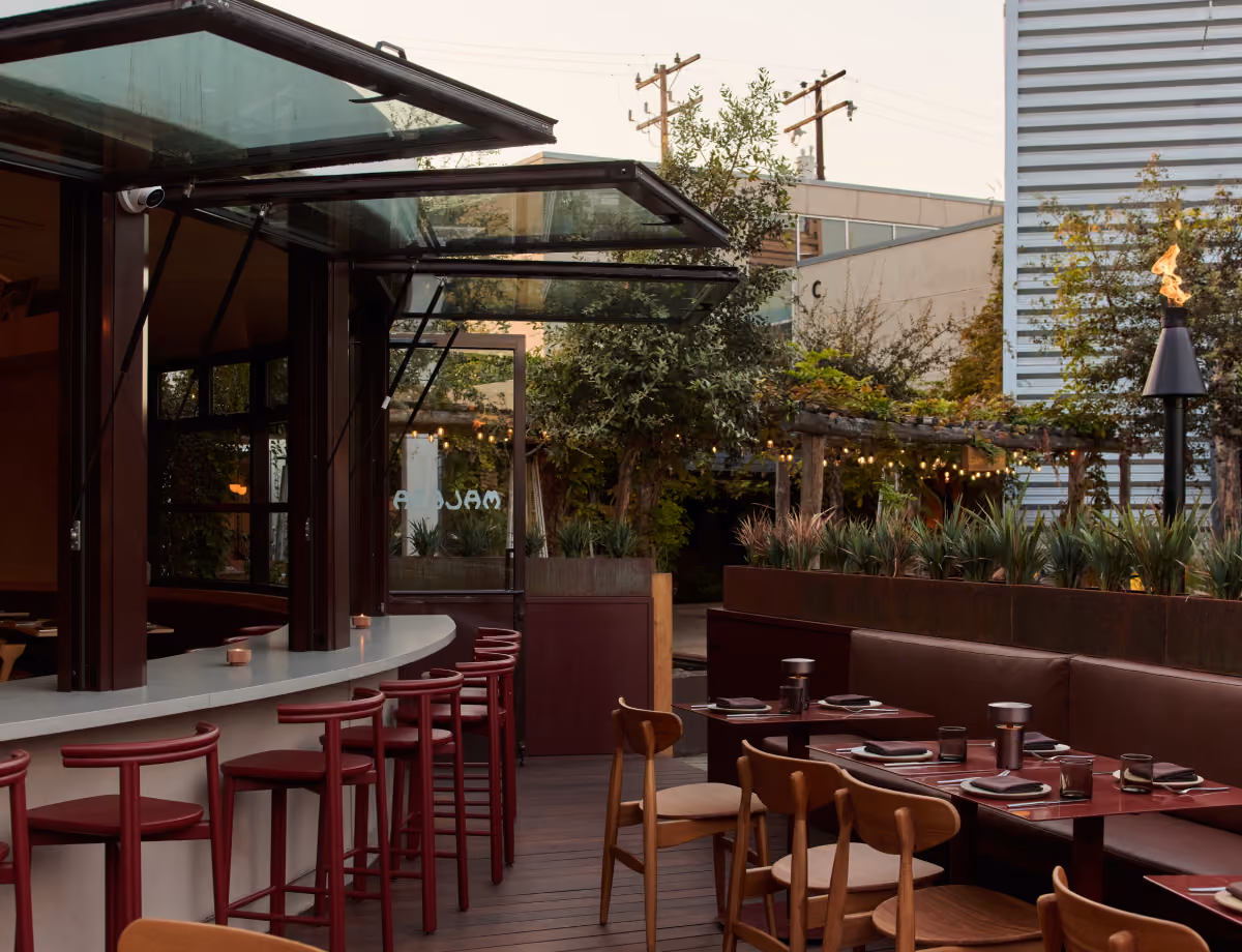 Outdoor patio with a bar lined with red stools and wooden dining tables under glass awnings and string lights.