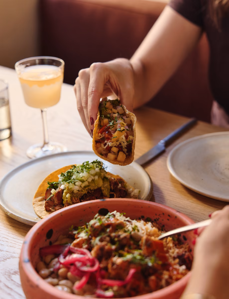A hand holds a taco above a plate of tacos and a bowl of rice with pickled onions, beside a pale citrus cocktail.