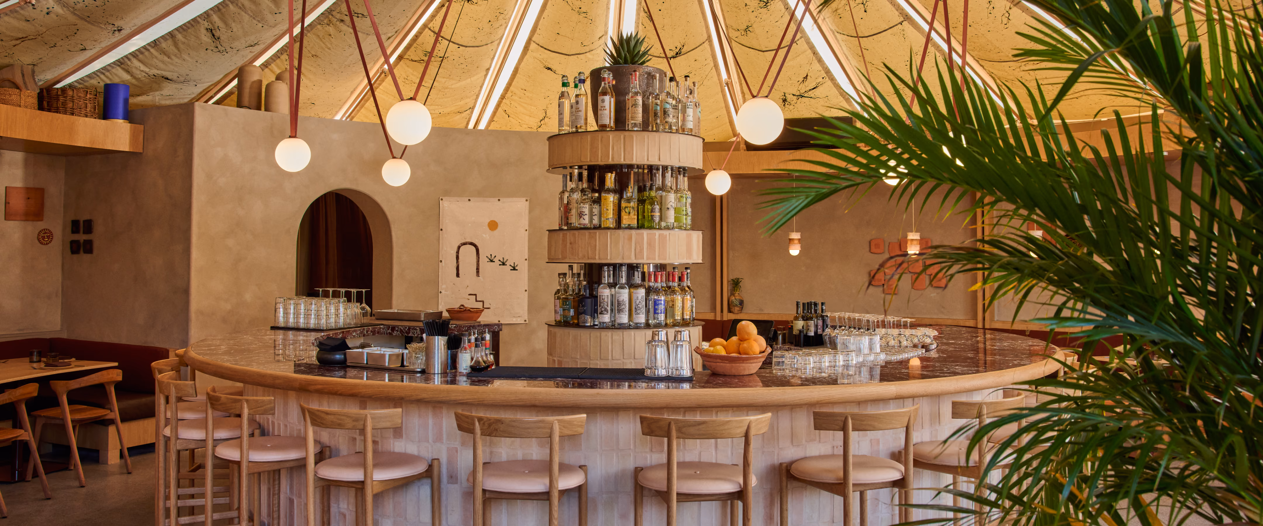 Curved bar at Maleza Costa Mesa with wooden stools, tiered bottle shelves and globe pendant lights under a conical ceiling, framed by palm fronds.