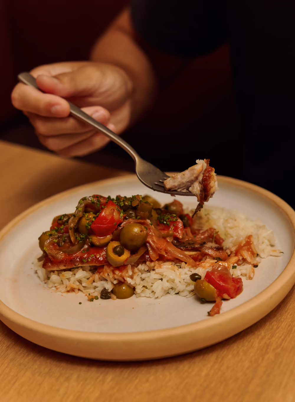 A hand lifts a forkful of shredded meat with olives and roasted peppers from a plate of rice.