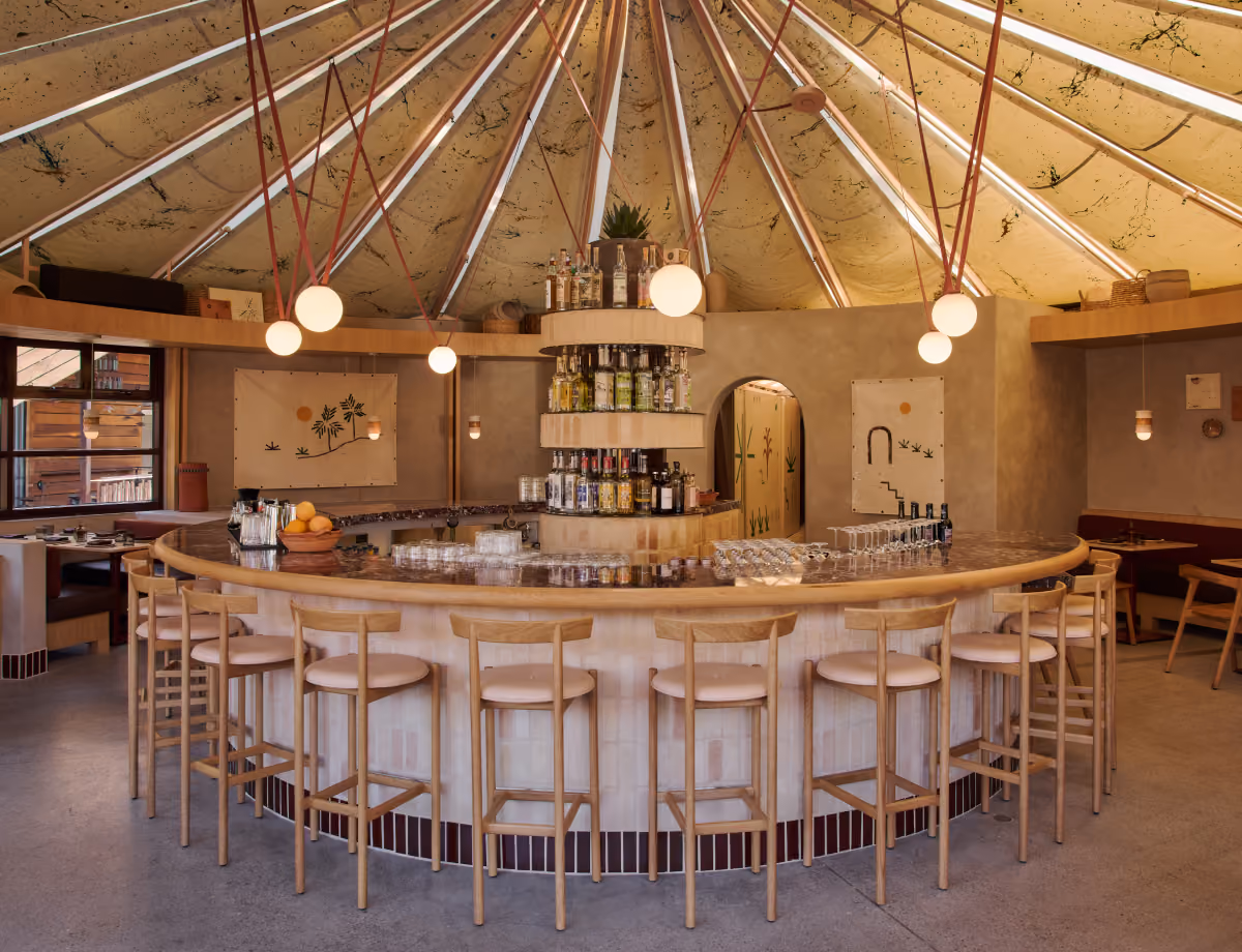 Curved bar with wooden stools beneath a tented conical ceiling, globe pendant lights and a tiered bottle display.