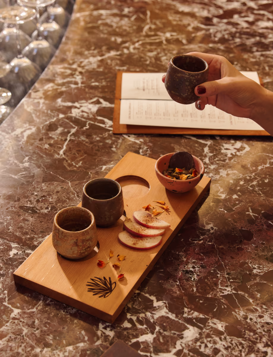 Mezcal tasting flight in clay cups on a wooden board with orange slices and chile salt, on a marble bar.