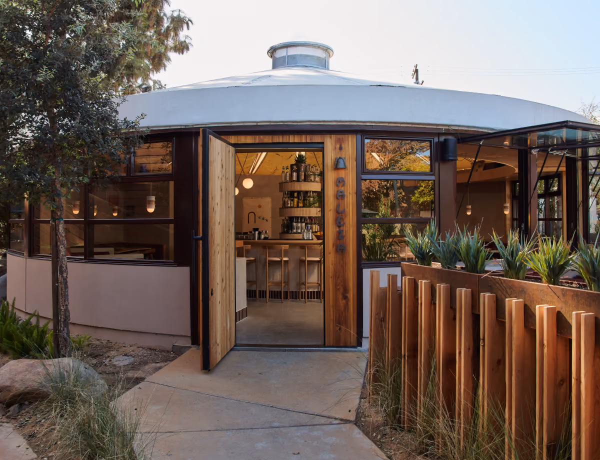 Exterior of Maleza Costa Mesa, a round building with an open wooden door, surrounded by trees, agave and a wooden fence.