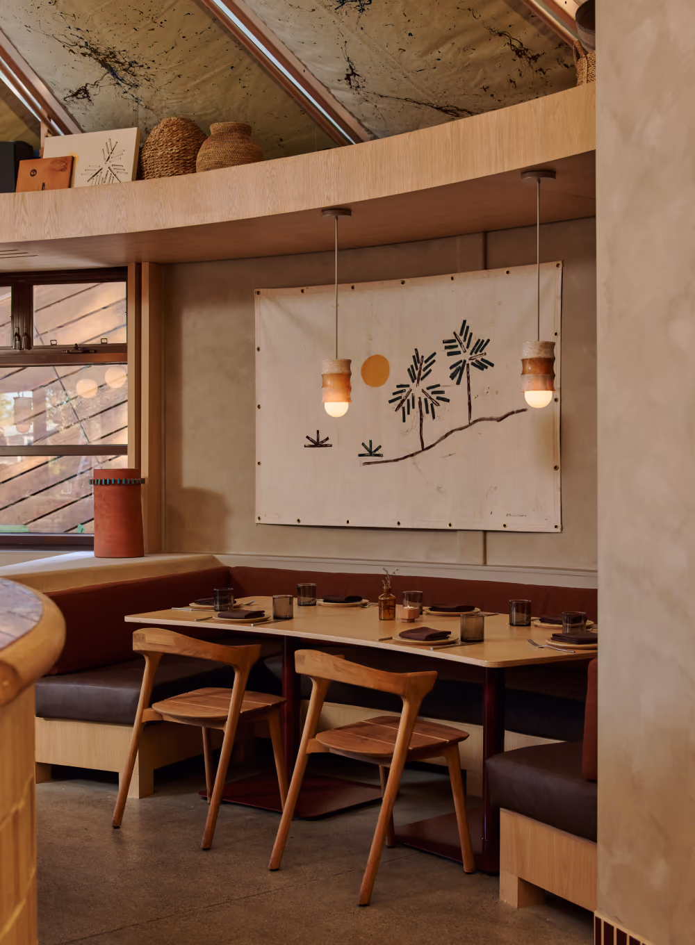 Booth dining nook at Maleza Costa Mesa with wooden chairs, pendant lights and a palm tree wall hanging beneath a curved ceiling.