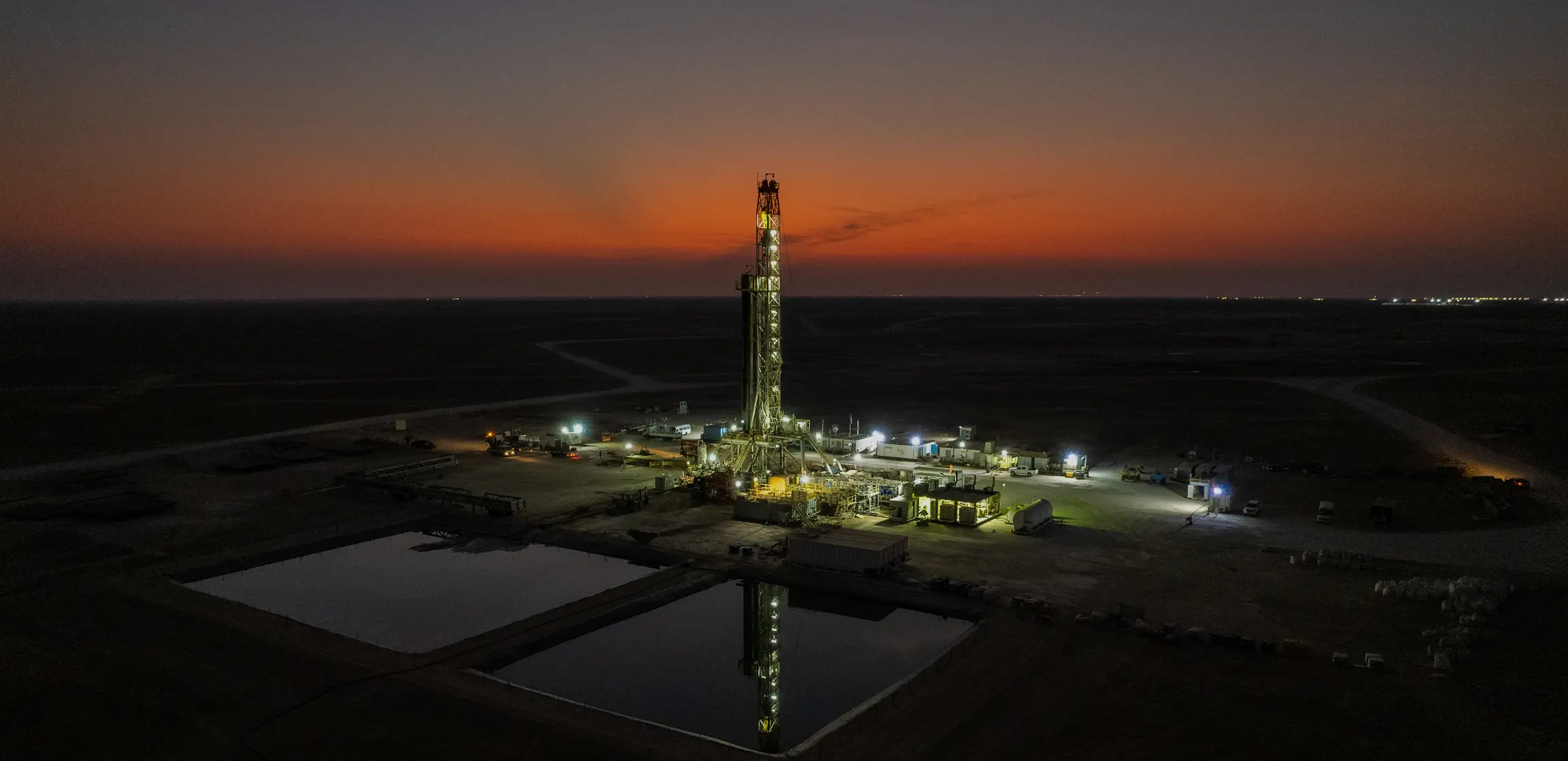 Aerial wide shot of an illuminated oil drilling rig at twilight in a flat desert landscape with reflection pools in the foreground.