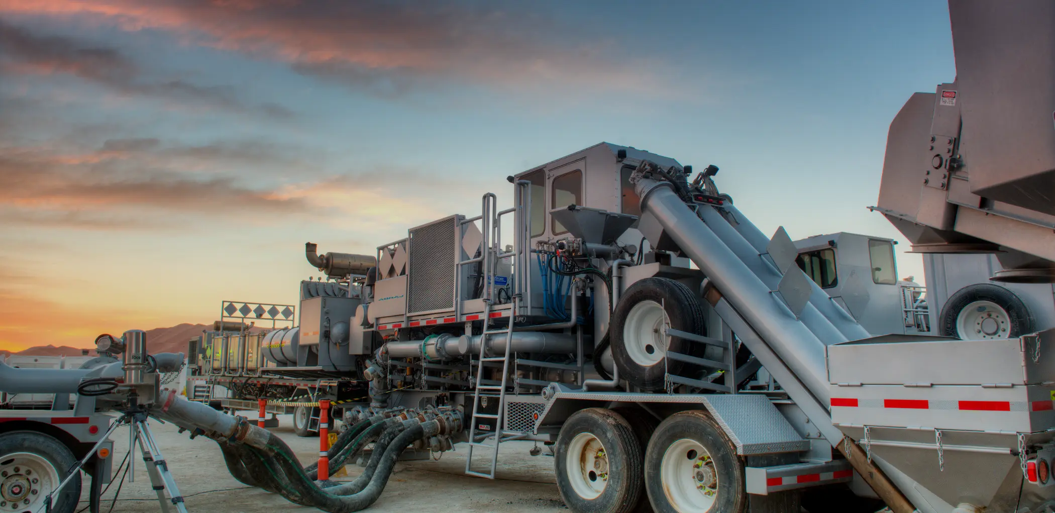 A heavy-duty hydraulic fracturing blender unit on a trailer at sunset, featuring large intake hoses and mixing equipment.