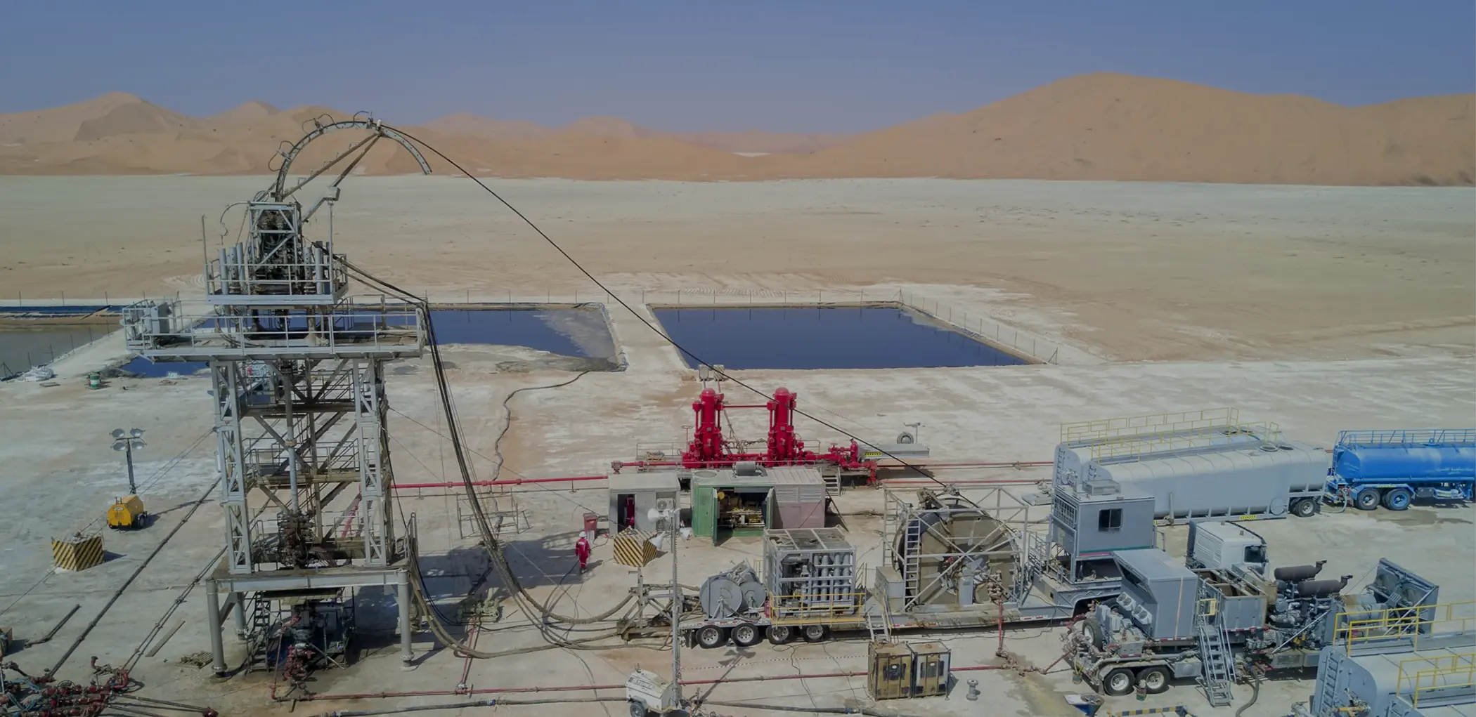 An aerial panoramic view of a large oilfield stimulation site in a desert with a coiled tubing unit, red pump manifolds, and fluid storage ponds.