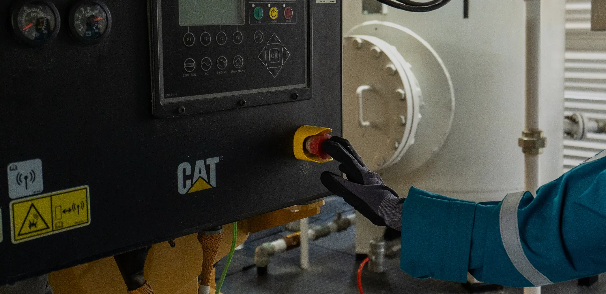 Close-up of a gloved hand pressing a red emergency stop button on a CAT industrial control panel.