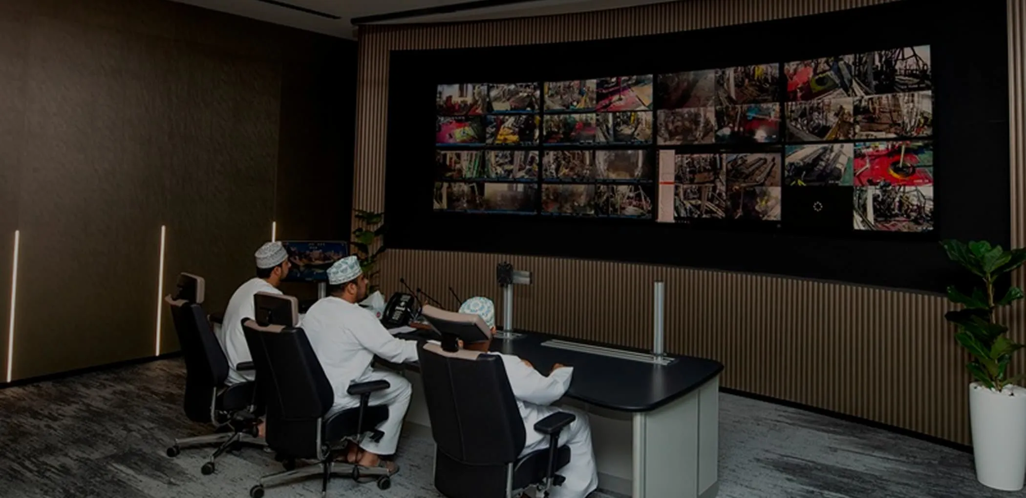 Three operators sitting at a sleek control desk in a modern command center, monitoring a large wall of video surveillance screens showing industrial operations.