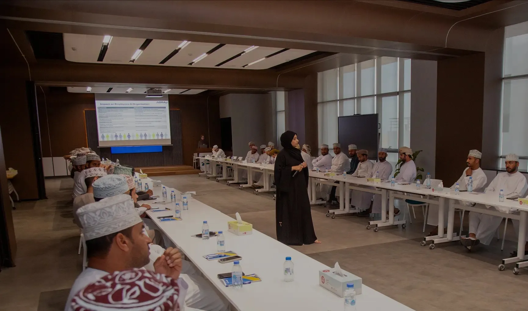A female instructor in a black abaya leading a corporate presentation for a group of men in traditional Omani dress in a modern, well-lit conference room.