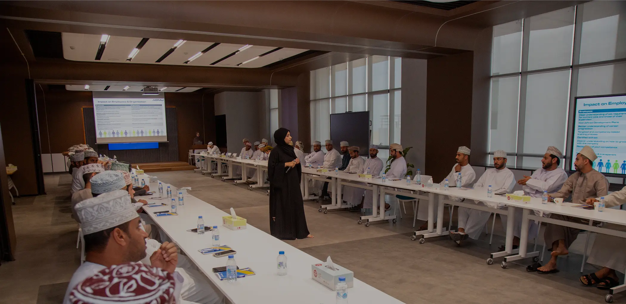 A female instructor in a black abaya leading a corporate presentation for a group of men in traditional Omani dress in a modern, well-lit conference room.
