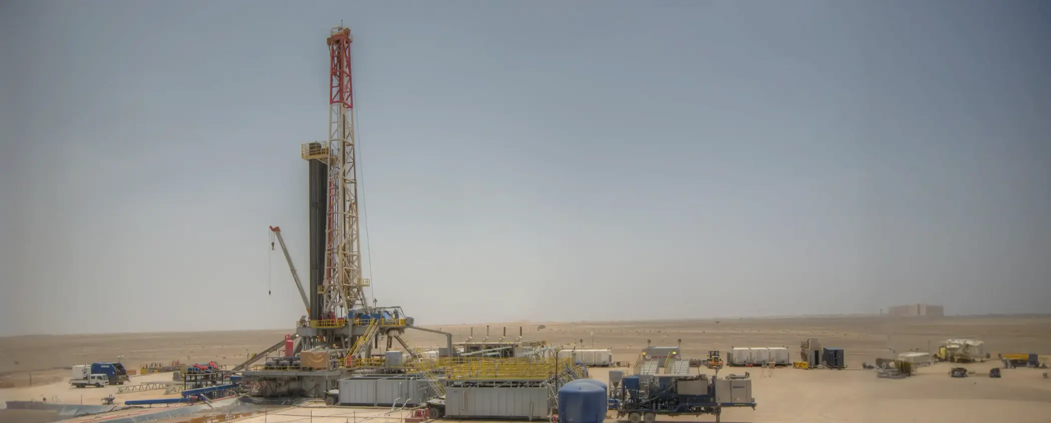 Wide panoramic shot of a land-based drilling rig and modular support units in a vast, arid desert under a clear sky.