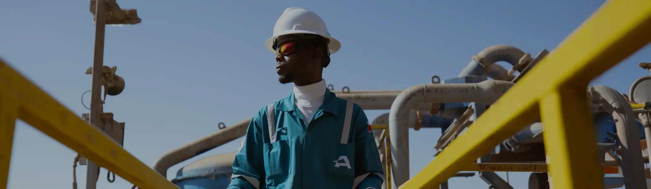 A worker in a turquoise jumpsuit and white hard hat standing on a yellow industrial walkway against a clear sky.