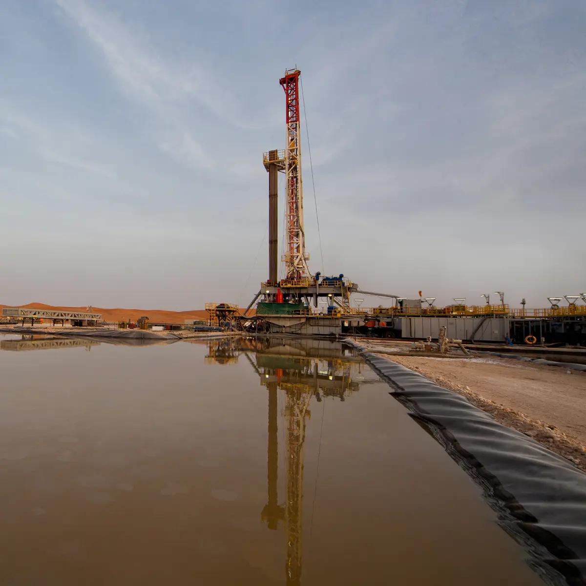 A tall drilling rig reflected in the calm surface of a large, lined fluid storage pit at a desert well site during dusk.
