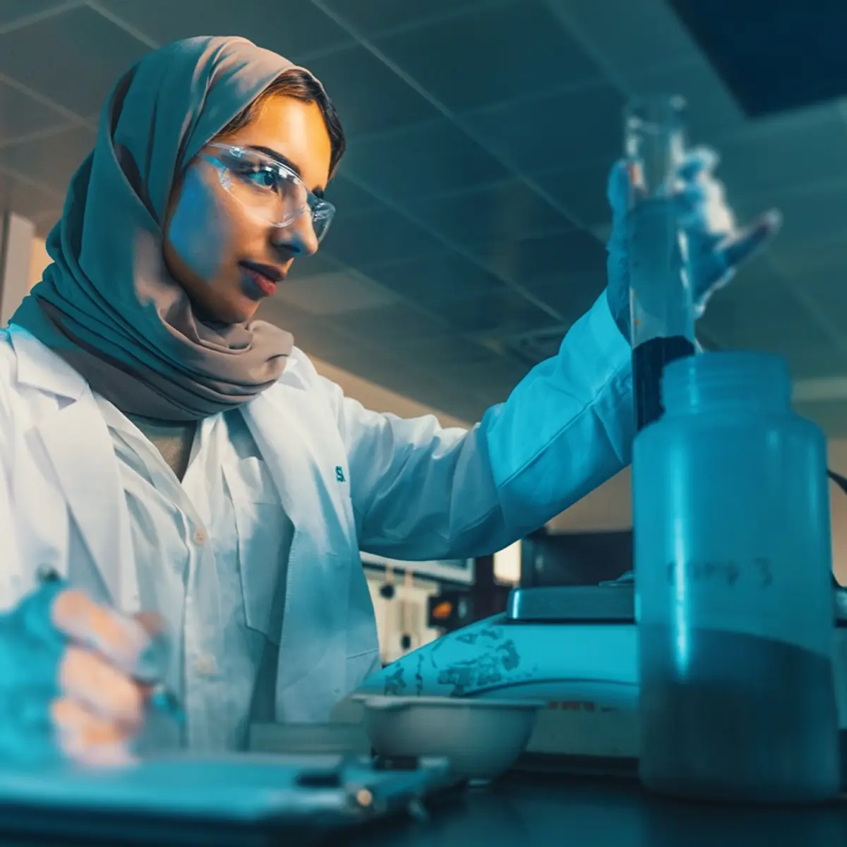 A female chemist wearing a hijab, lab coat, and safety glasses conducting an experiment with a graduated cylinder in a laboratory.