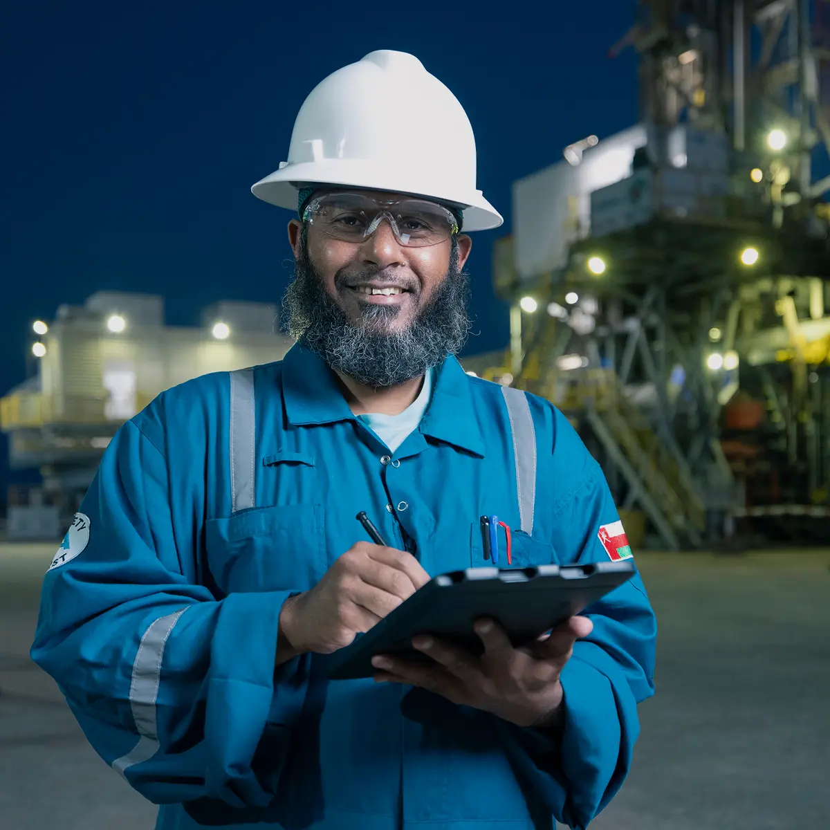 Portrait of a smiling oilfield engineer in a blue jumpsuit and safety gear holding a digital tablet at night with a rig in the background.