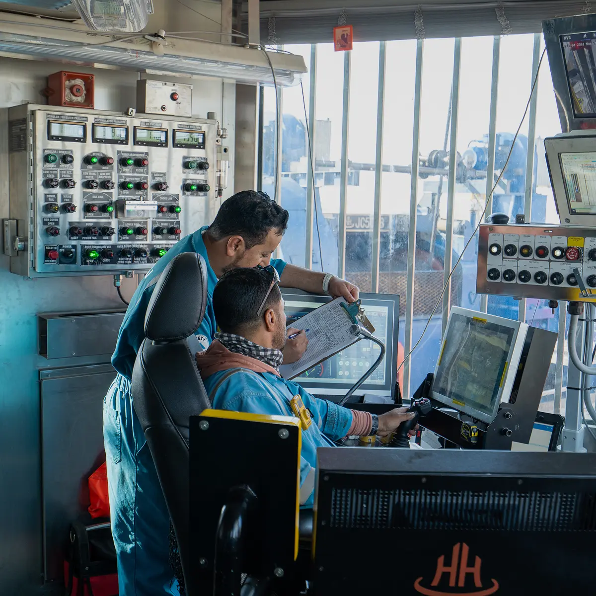 Two technicians in blue coveralls working together inside a control room, reviewing a checklist against a wall of industrial gauges and computer monitors.