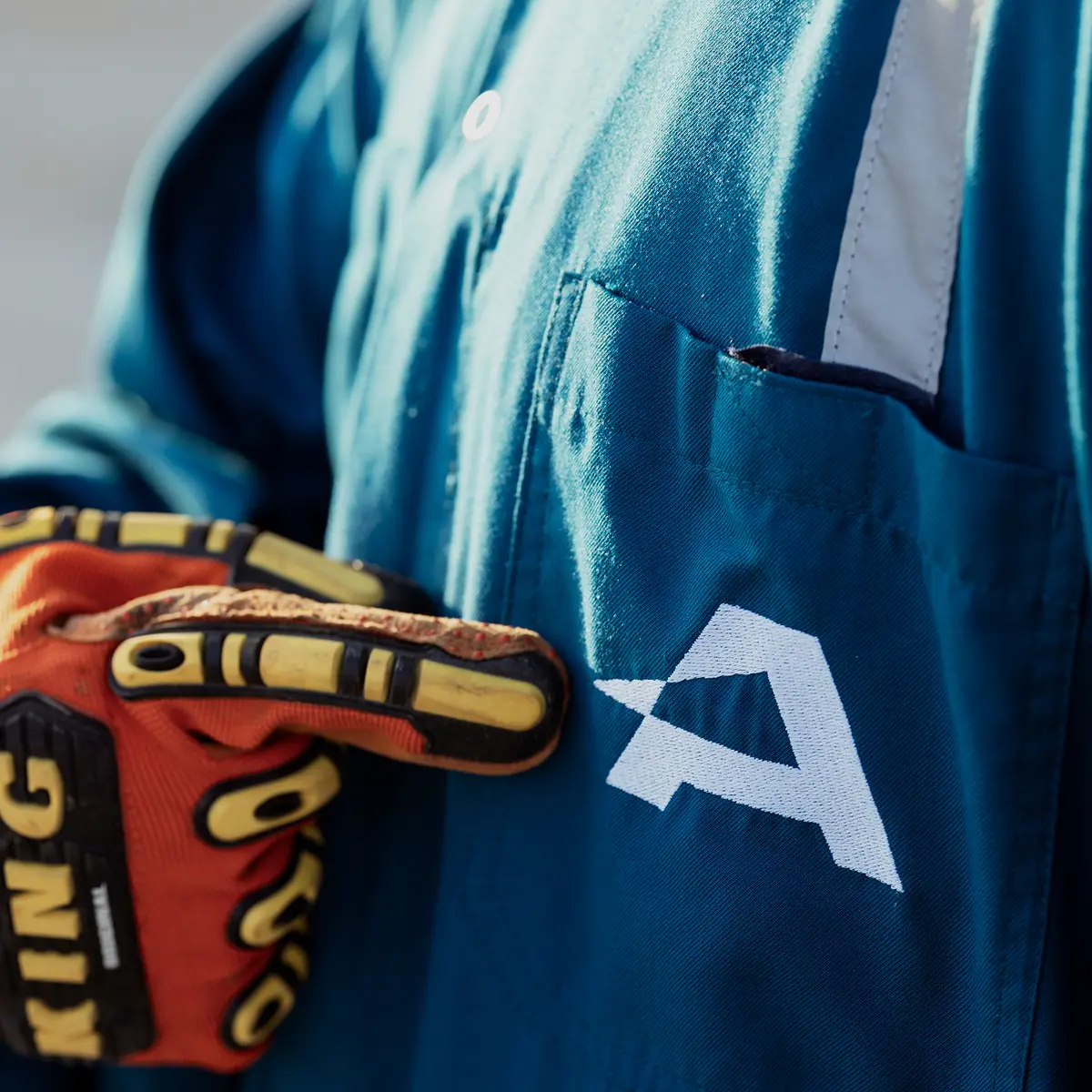 Close-up of a worker's blue industrial jumpsuit featuring the white Abraj company logo, with a hand in an orange safety glove pointing to the emblem.