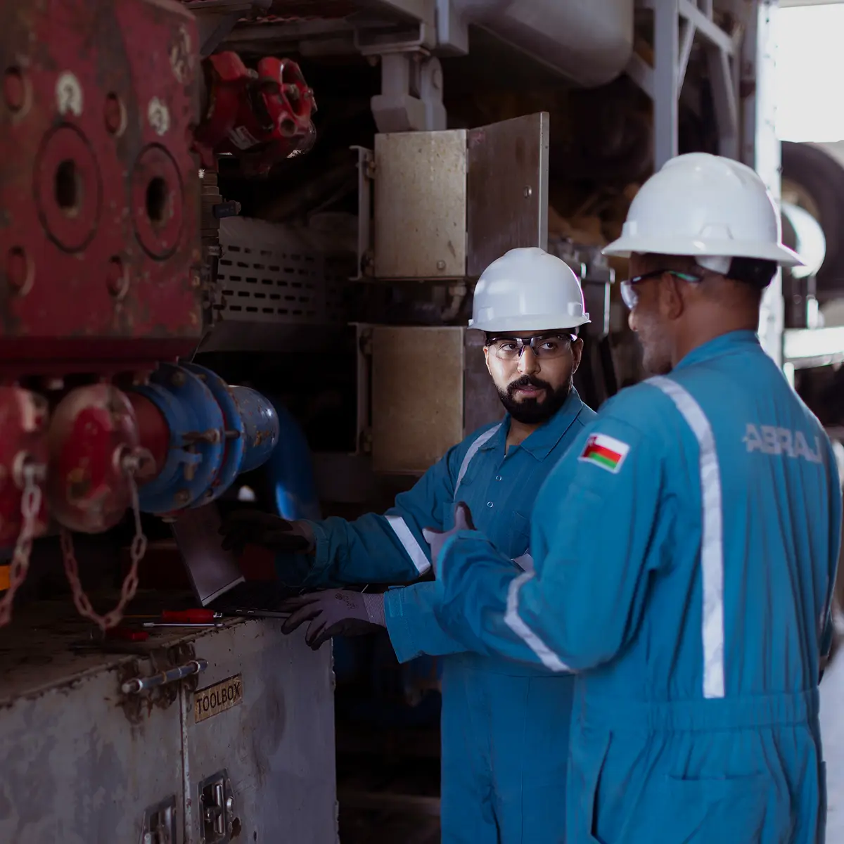 Two field engineers in blue coveralls and white hard hats using a laptop to troubleshoot heavy machinery at an outdoor industrial site.