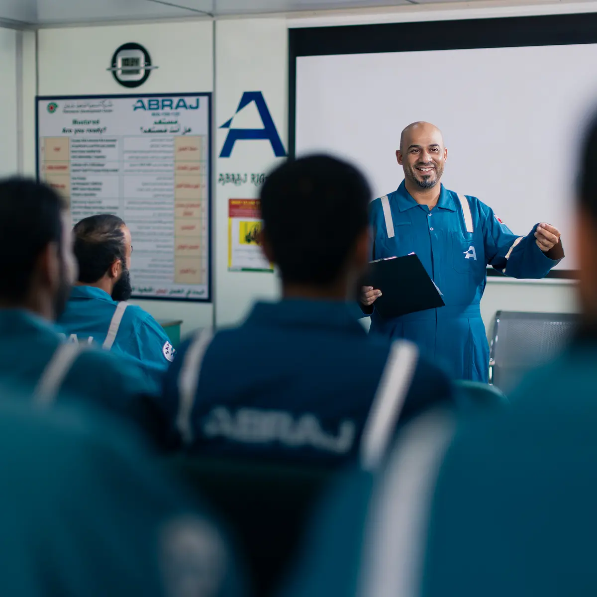 A smiling instructor in a teal jumpsuit standing at the front of a training room, holding a clipboard and addressing a group of seated employees.