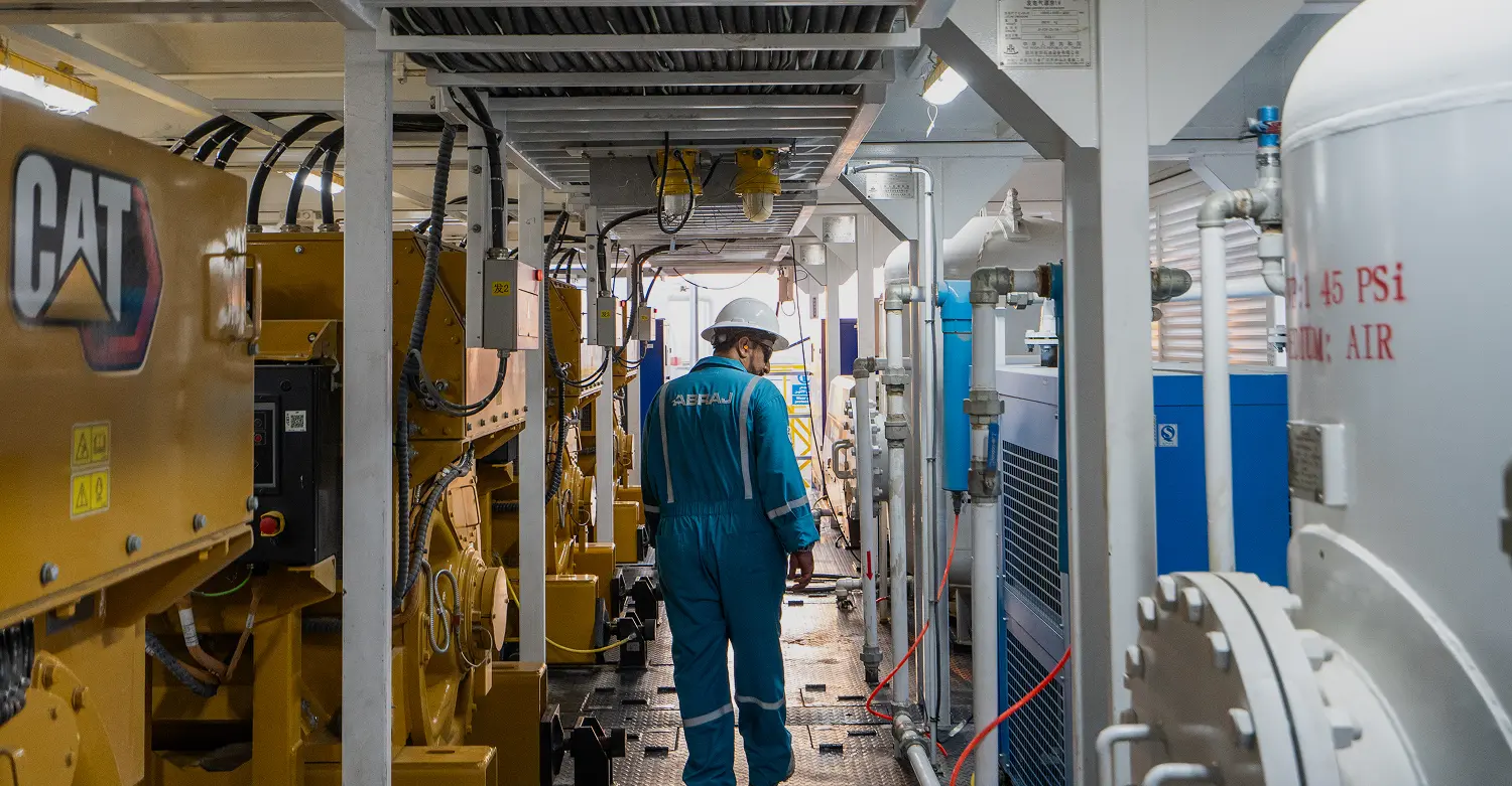 An engineer in a blue jumpsuit and white hard hat walking through a technical room filled with CAT machinery and piping.
