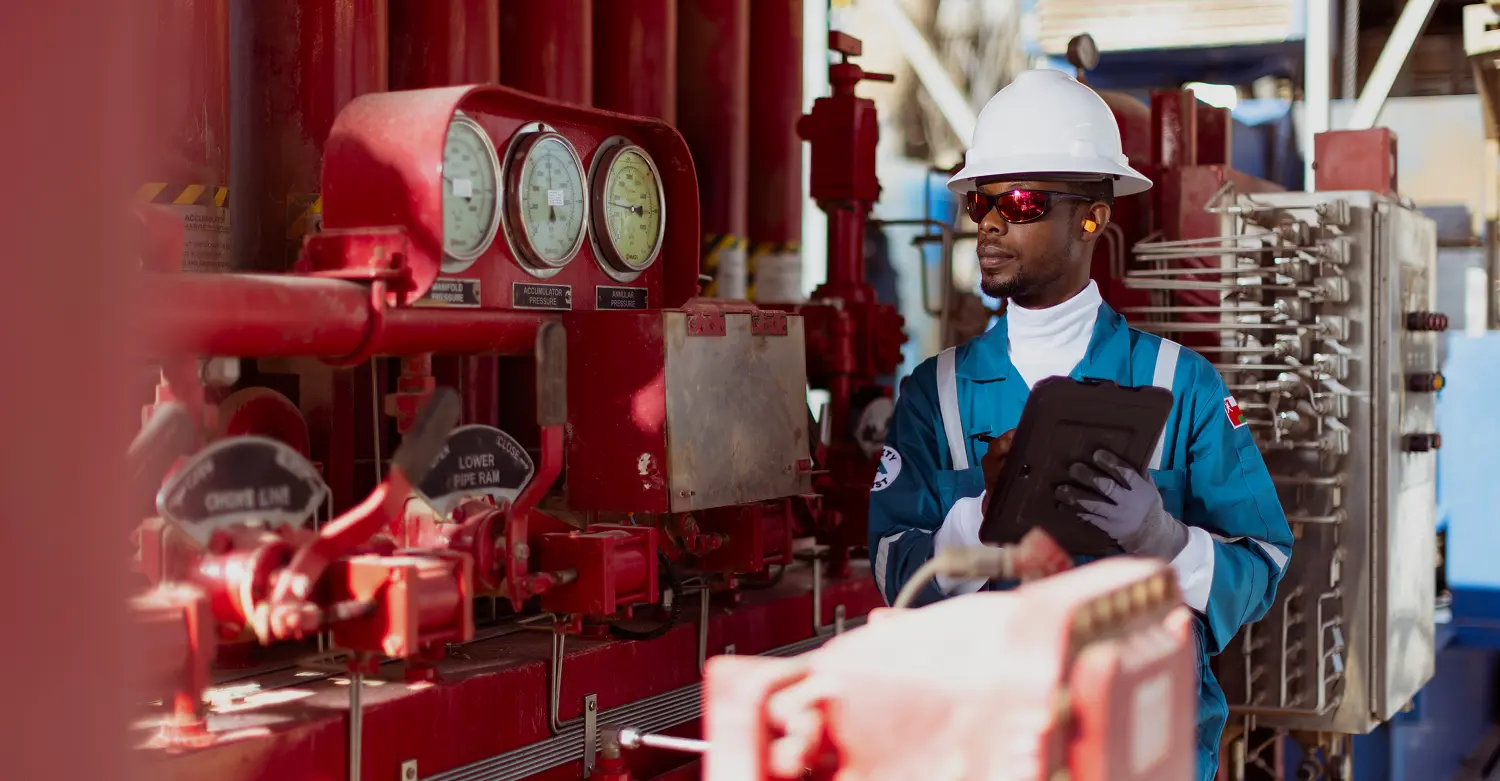 A technician in a blue jumpsuit and white hard hat using a tablet to monitor data next to a red control panel with pressure gauges.