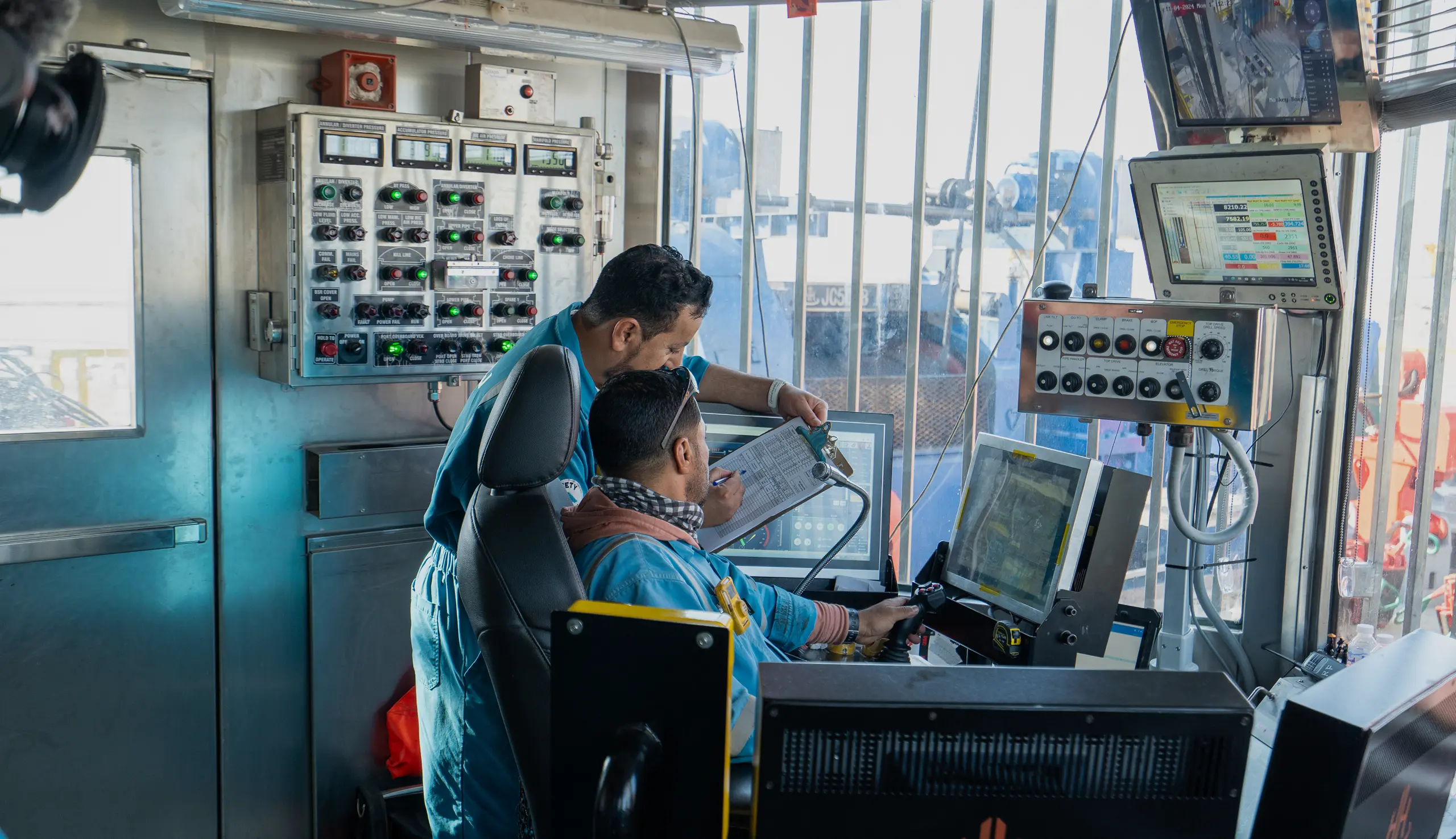 Two technicians in blue uniforms reviewing data on a clipboard and digital monitors inside an industrial control cabin.