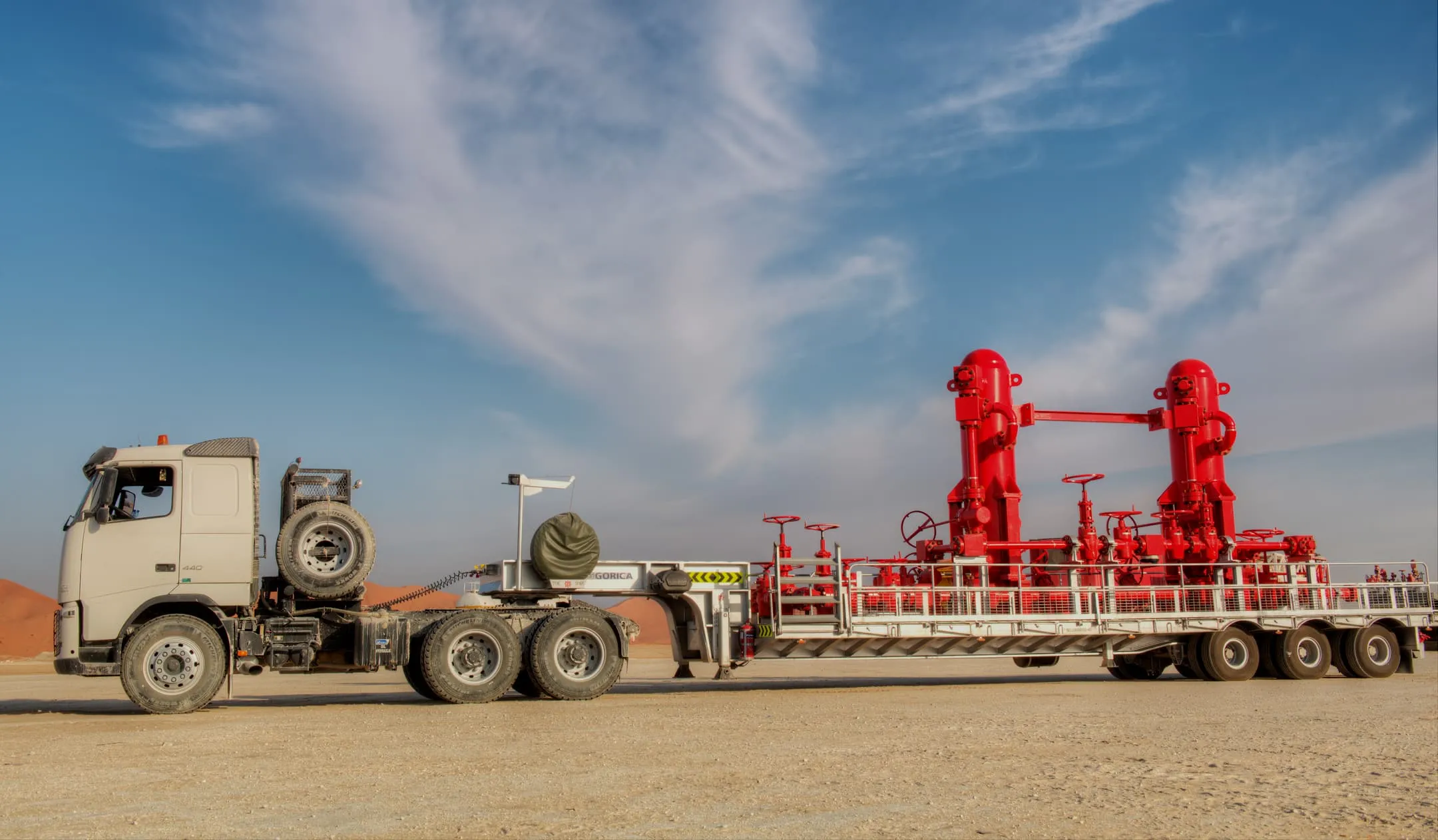 A white heavy-duty truck hauling a trailer with red industrial testing vessels through a desert environment under a blue sky.