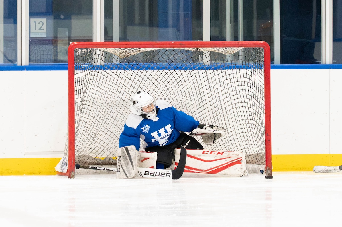 Ice hockey goalie in blue jersey and white helmet crouching in front of goal net on ice rink.