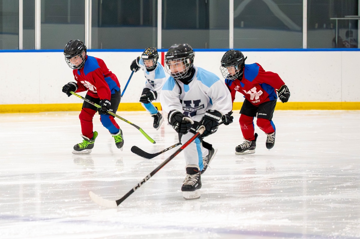 Four young ice hockey players wearing helmets and skates, two in red and two in white jerseys, skating on ice rink during a game.