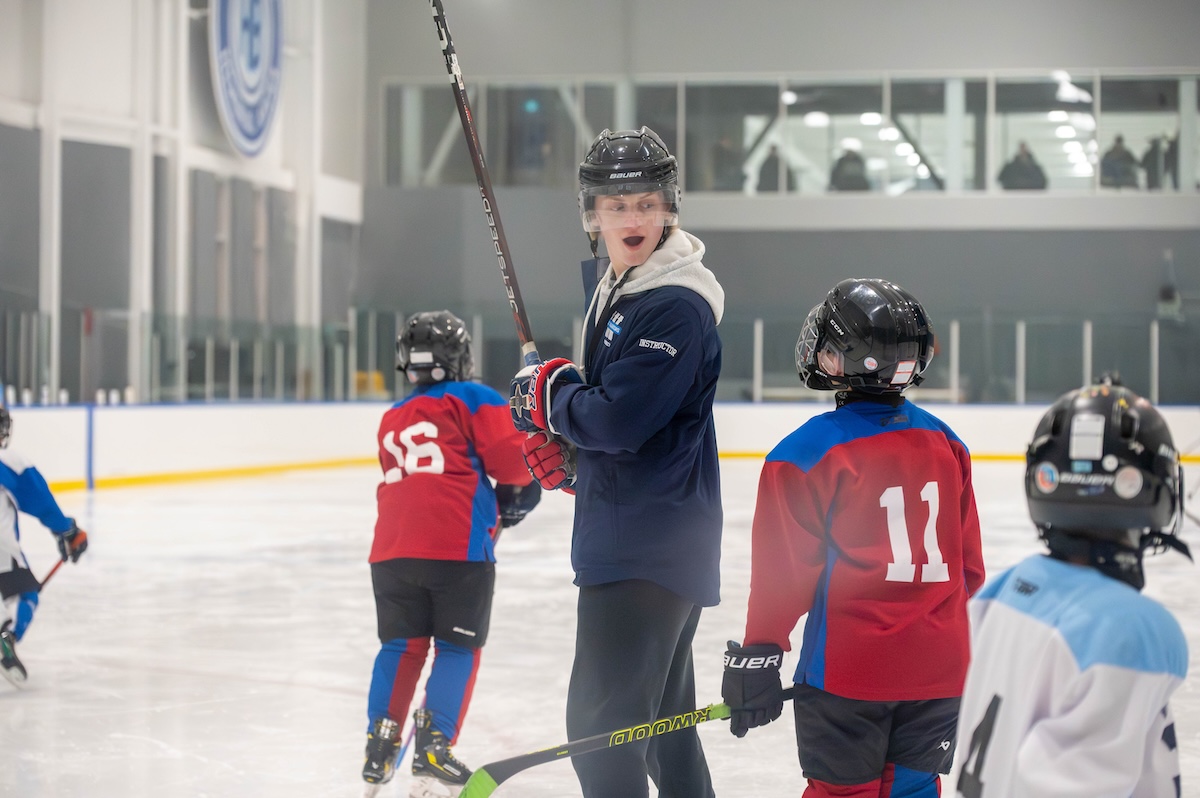 Hockey instructor wearing a helmet and gloves talks to young players on ice rink during practice.