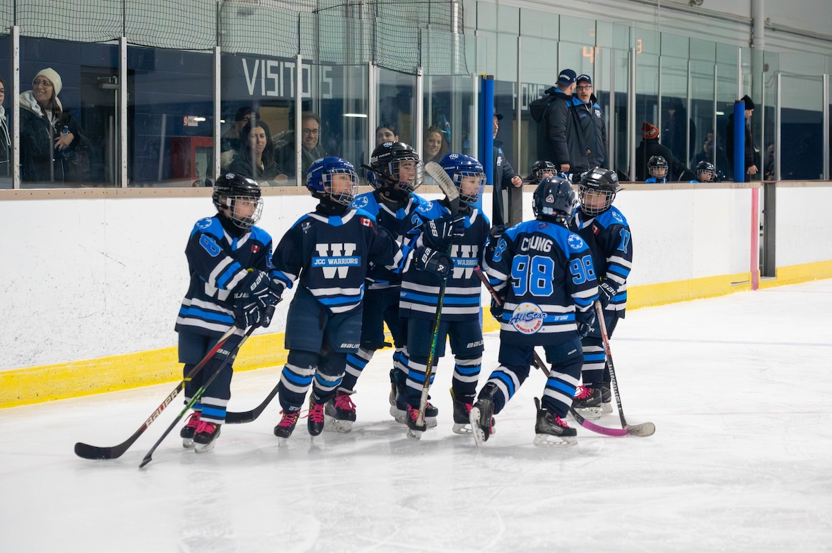 Group of young hockey players in blue JCC Warriors jerseys standing together on an ice rink during a game.