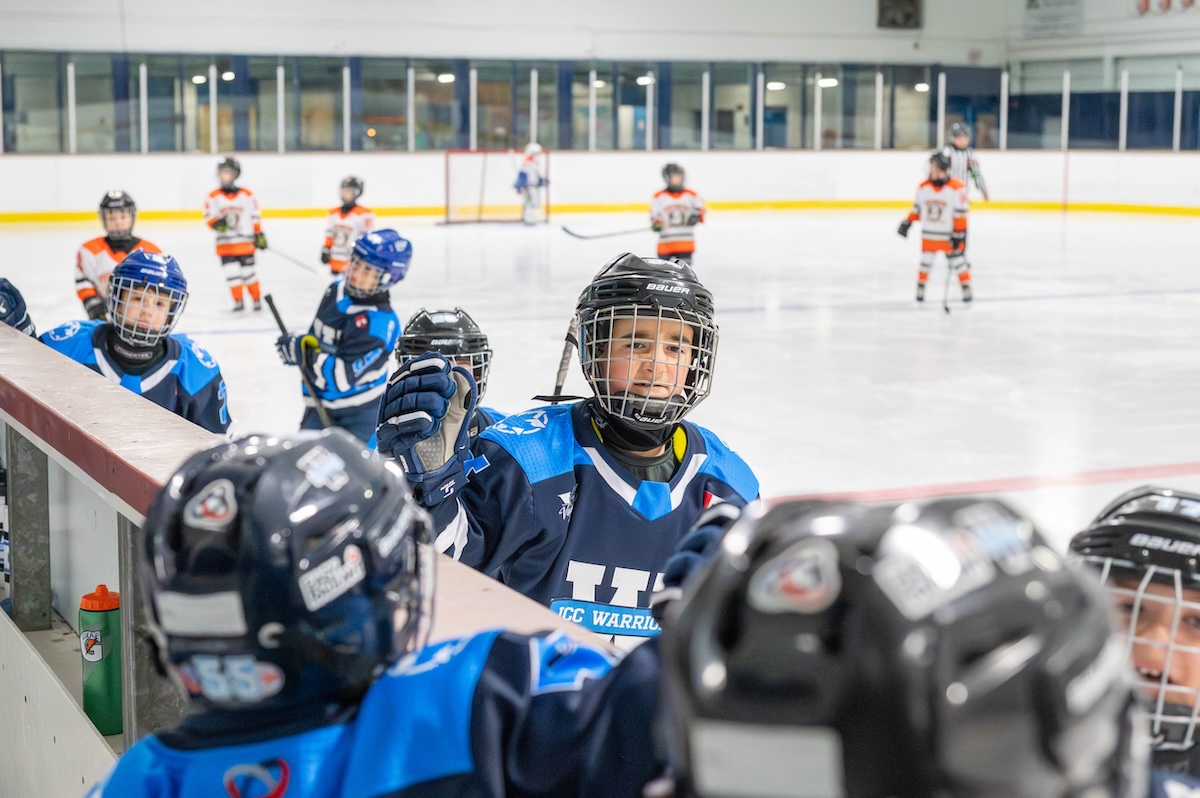 Youth ice hockey players in blue and black uniforms giving high-fives near the rink boards with players in white and orange uniforms visible in the background.
