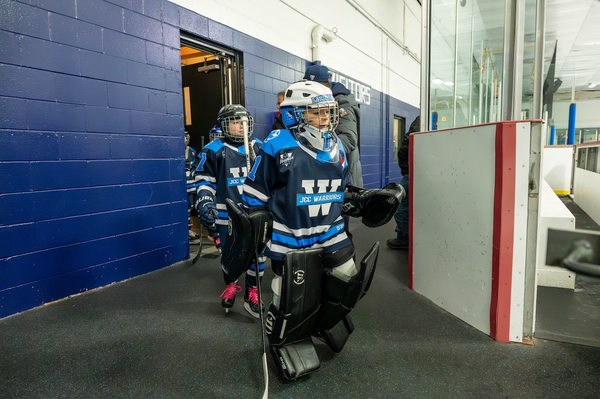 Youth hockey players in blue JCC Warriors uniforms walking out from locker room onto ice rink.