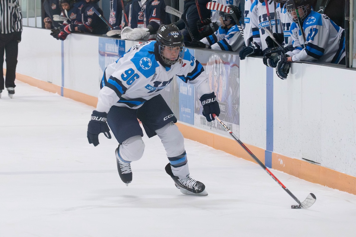 Ice hockey player in white and blue jersey skating with the puck near the rink boards while teammates watch.