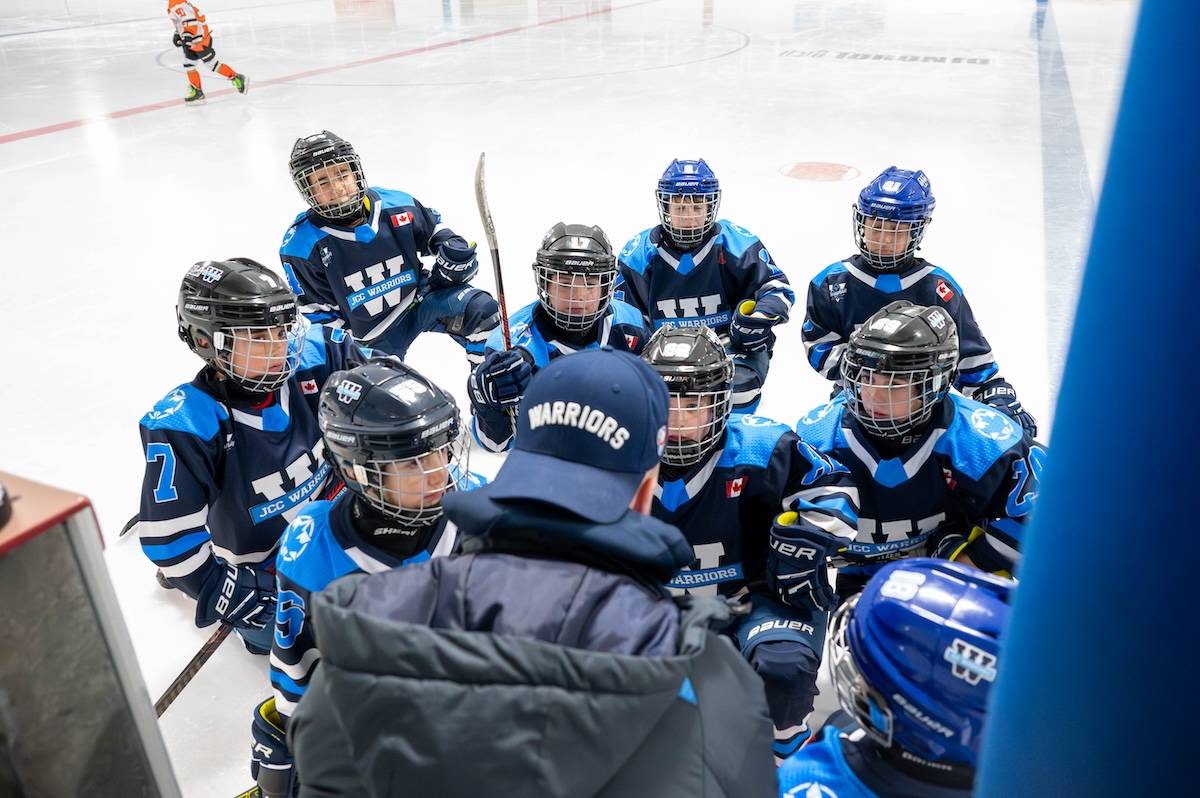 Youth ice hockey team in blue uniforms listening to coach during a rink timeout.