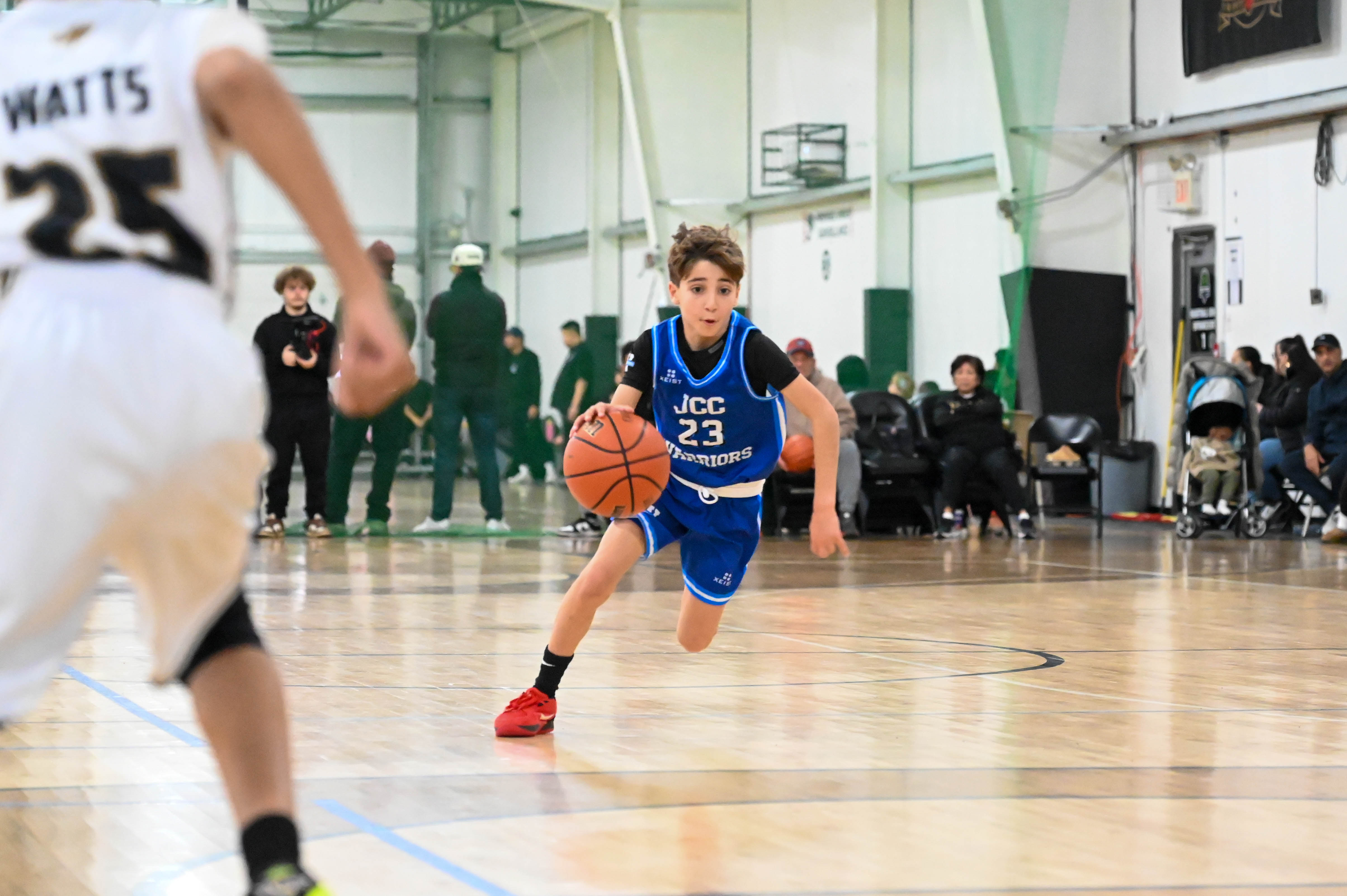 Youth basketball player in blue JCC Warriors uniform dribbling the ball on an indoor court during a game.