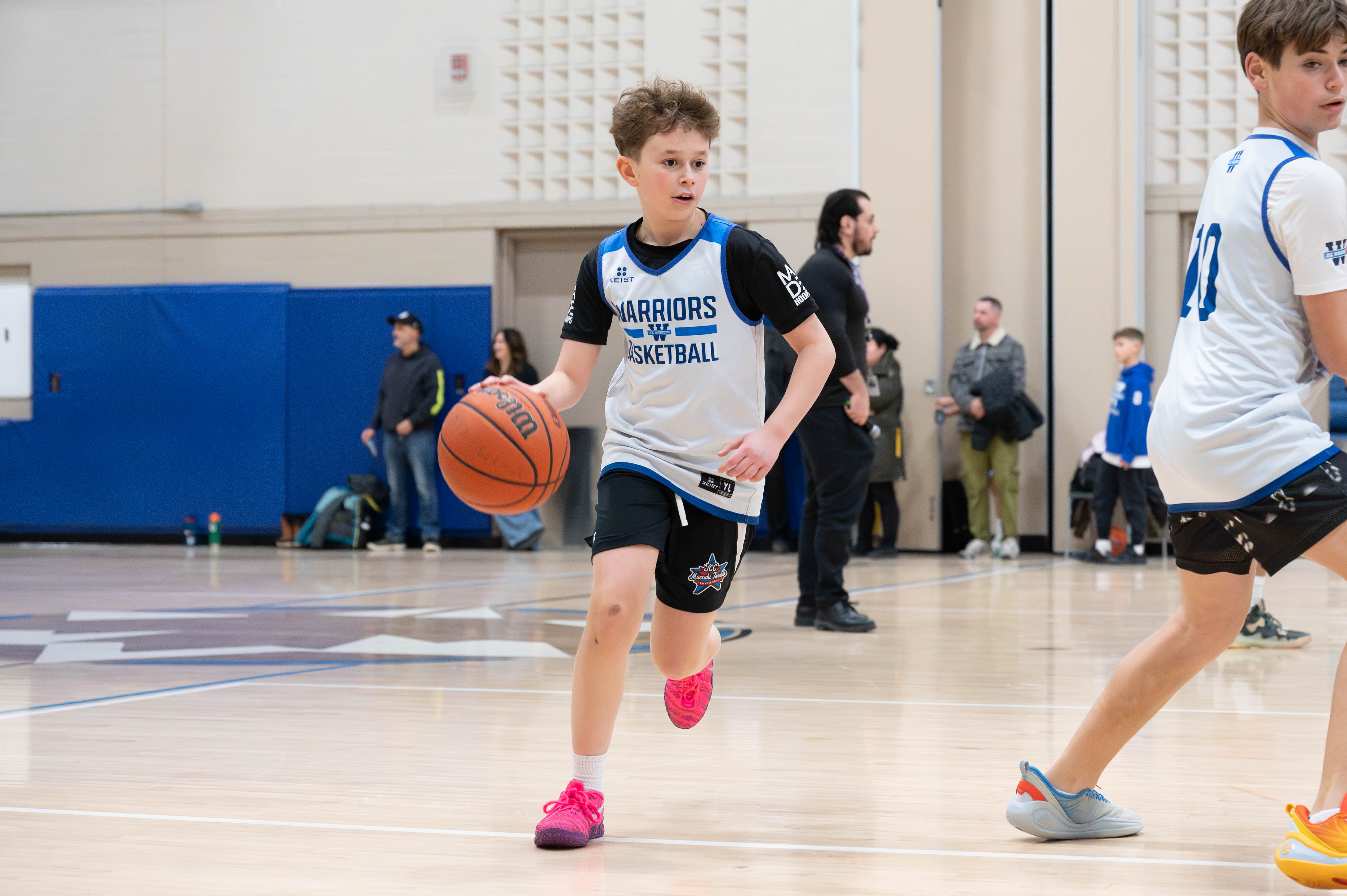 Young boy in Warriors Basketball jersey dribbling a basketball on an indoor court during a game.