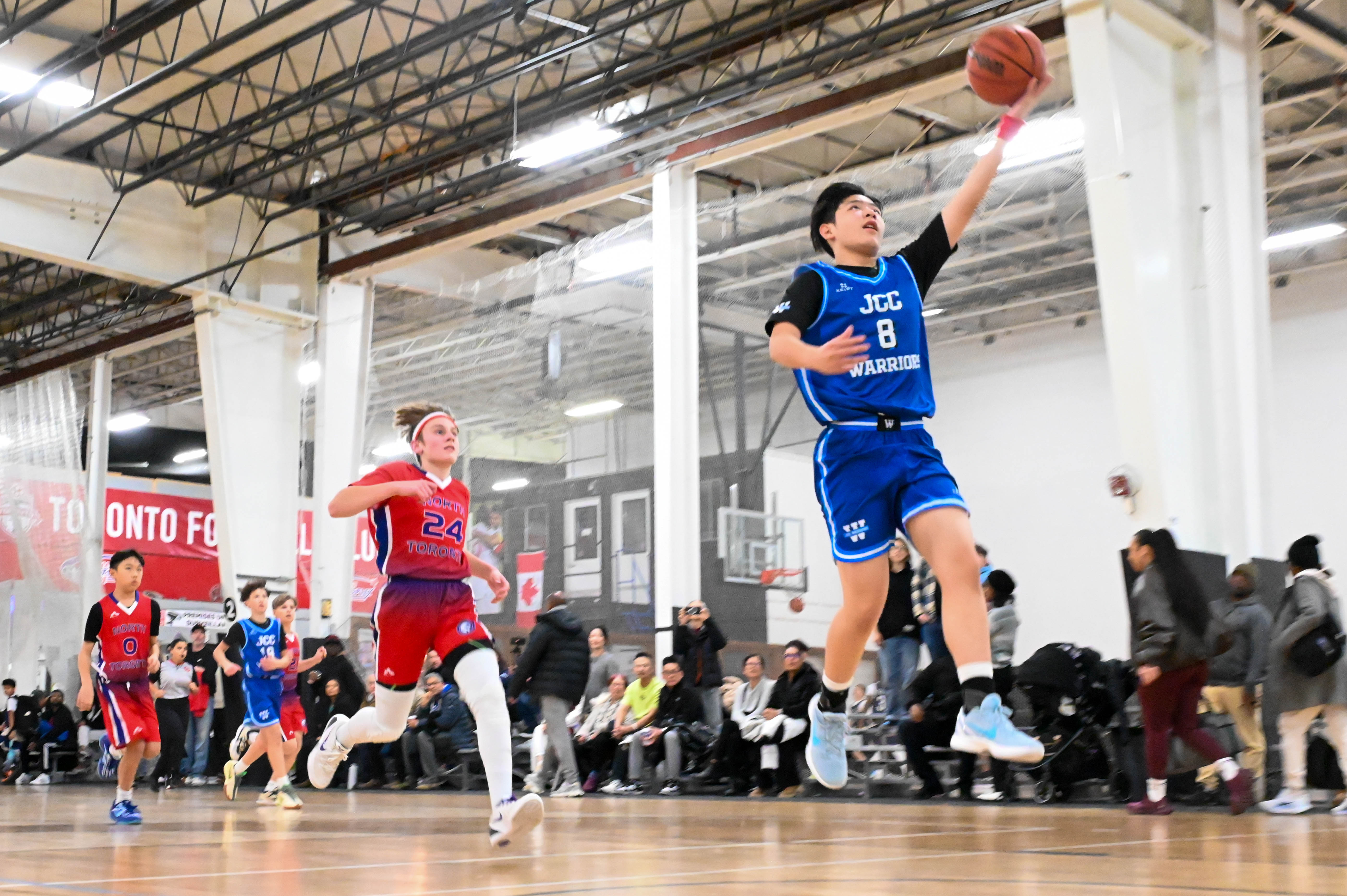 Young basketball player in blue jersey jumping to make a layup while a player in red jersey runs behind.