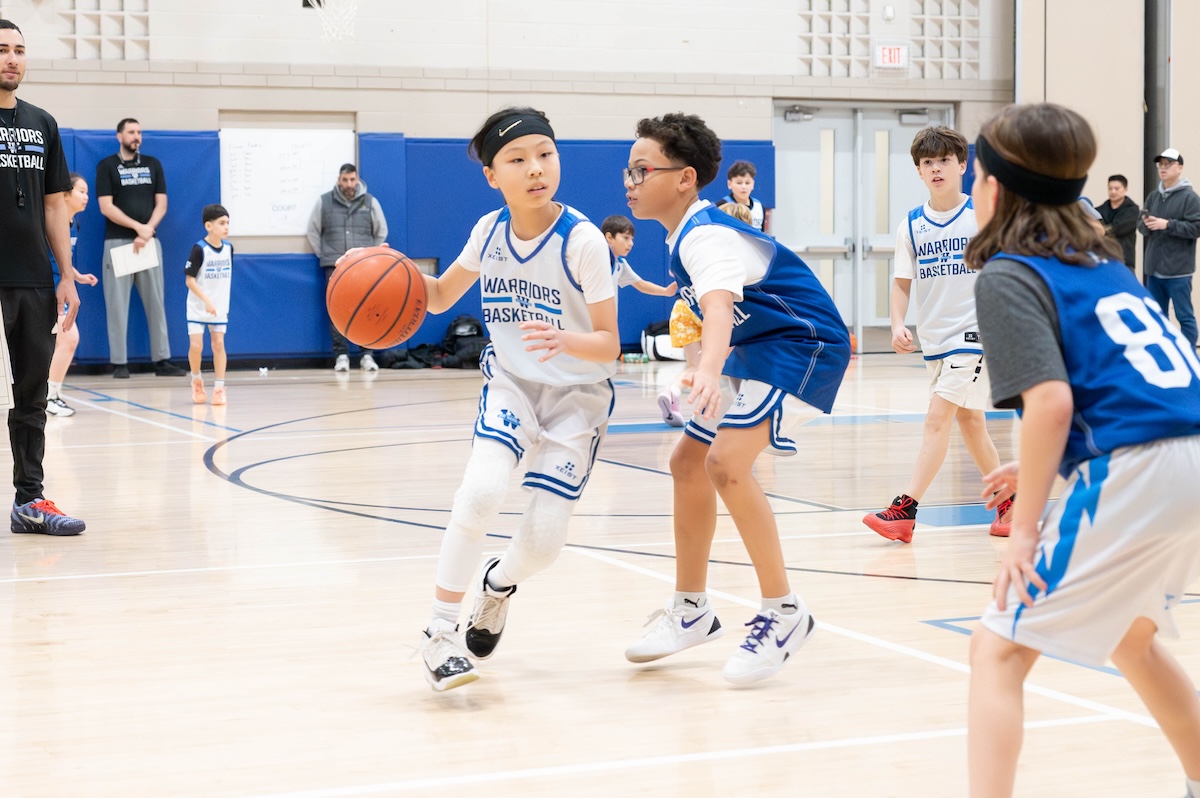 Youth basketball players in white and blue Warriors jerseys playing on indoor court with coaches and spectators in background.