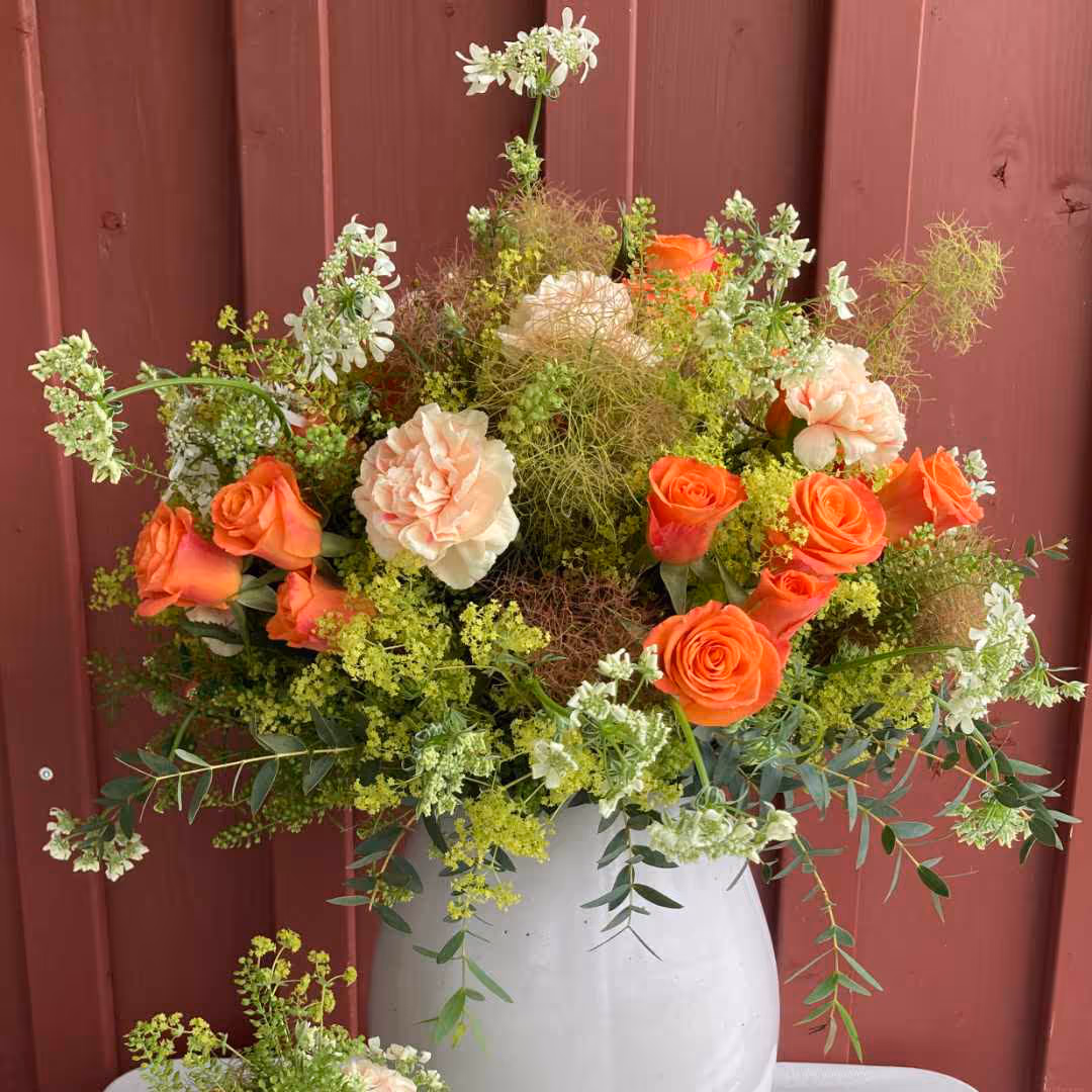 Blumenstrauß mit orangefarbenen Rosen, rosa Nelken und grünen, gelben sowie weißen Blumen in einer weißen Vase vor roter Holzwand.